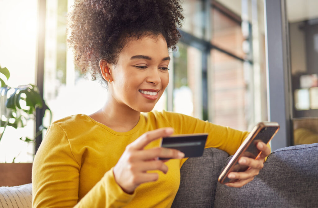 Una mujer negra con una blusa amarilla está sentada en el sofá con una tarjeta de crédito en una mano y su teléfono celular en la otra. Ella está aprendiendo las mejores tarjetas de crédito para ganar millas.