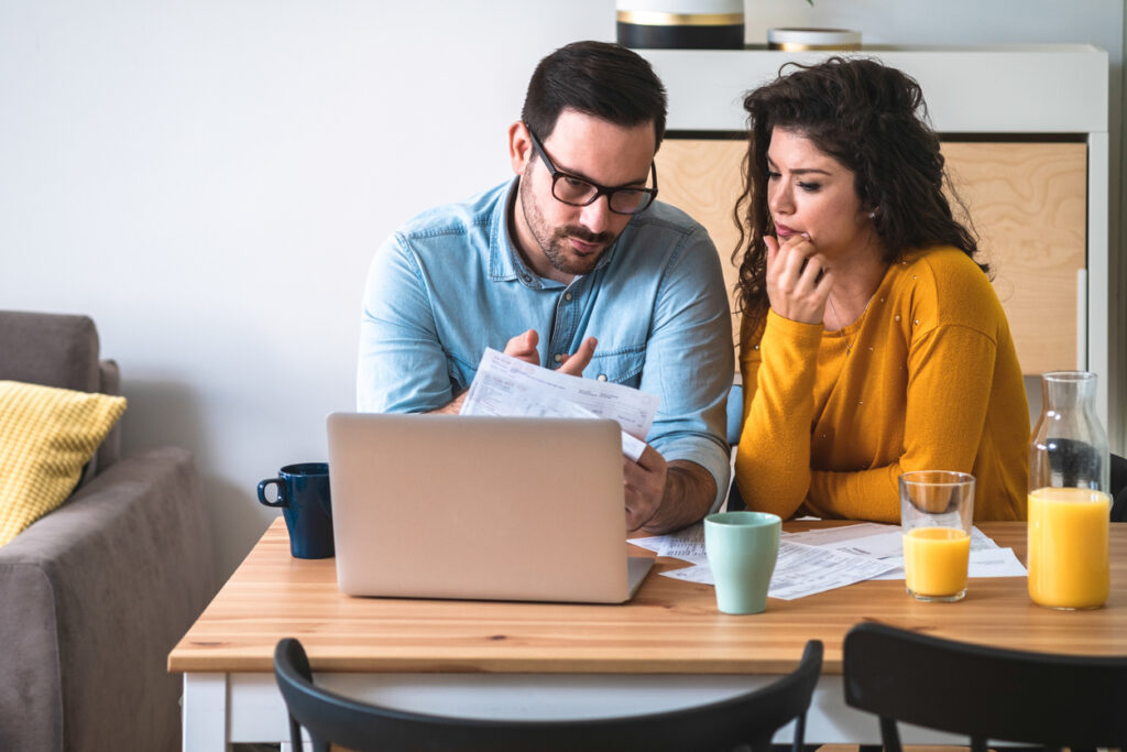 Una pareja está sentada en una mesa que contiene una computadora, una taza de café y algunos cafés. El hombre sostiene una hoja de papel y le explica a su esposa cómo obtuvo ingresos extra vendiendo millas a MercoMillas