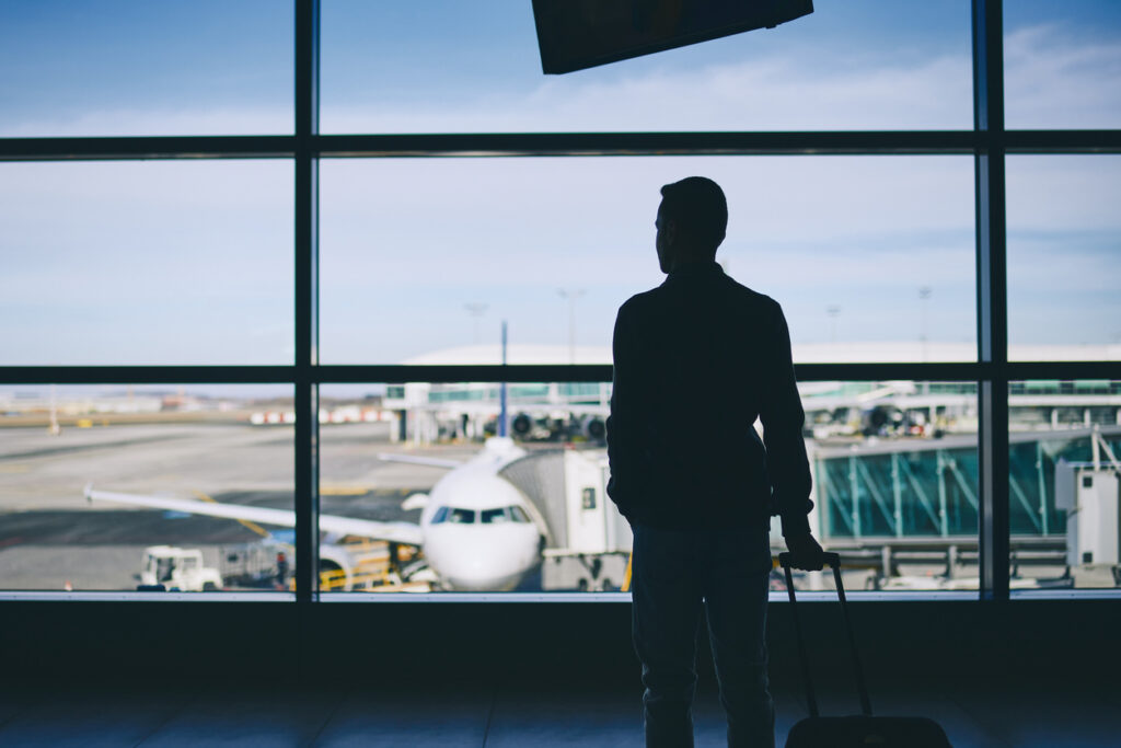 Un hombre está de pie en una sala de embarque sosteniendo su maleta mientras observa cómo el avión se prepara para despegar. Está aprovechando los beneficios de LATAM Pass para viajar más