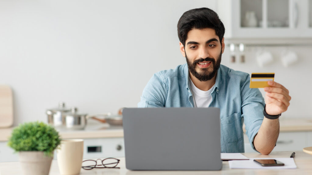 Un hombre blanco con cabello negro y barba negra, una chaqueta de mezclilla azul y una blusa blanca está sentado en una mesa en la cocina, frente a su cuaderno. Sostiene una tarjeta de crédito amarilla para verificar cuántos puntos acumuló para vender millas aéreas a MercoMillas