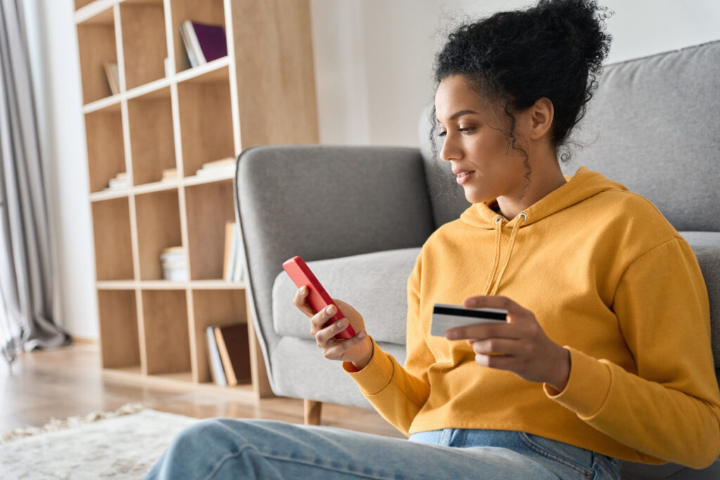 Una mujer morena con cabello negro rizado, blusa amarilla y jeans está sentada en el suelo, jugueteando con su teléfono celular. Con una mano sujeta el dispositivo y con la otra sujeta una tarjeta de crédito porque ha descubierto que esta es la mejor forma de acumular millas