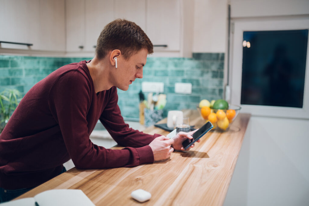 Un hombre blanco rubio con una blusa burdeos lleva auriculares mientras juguetea con su teléfono celular en la mesa. Tiene una tarjeta de crédito en la mano porque acaba de vender millas como una forma de ganar más dinero